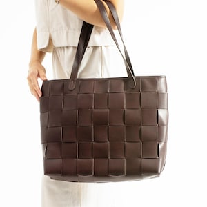 Può includere: Una borsa tote marrone scuro con un design intrecciato. La borsa ha due manici ed è tenuta da una persona. La trama della borsa è composta da blocchi quadrati. Lo sfondo è bianco.
