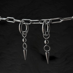 Op de afbeelding: Een zilverkleurige ketting met twee oorringen die aan de schakels zijn bevestigd. Aan elke ring hangt een kleine metalen kraal gevolgd door een scherpe, kegelvormige puntbedel. Het sieraad is volledig gemaakt van gepolijst metaal.