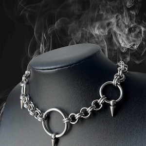 Puede incluir: Un collar plateado con diseño de cadena, con grandes colgantes circulares y detalles puntiagudos. El collar se exhibe en un maniquí negro. La palabra "MARAZM" está en la parte superior de la imagen.