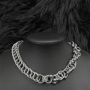 Puede incluir: Un collar de cota de malla plateado con una serie de anillos entrelazados y pequeños detalles de bolas. El collar se exhibe sobre un maniquí negro sobre un fondo oscuro y texturizado. La marca "DECHAINER" es visible.