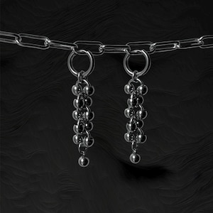 May include: A pair of silver-toned earrings with a chain and a cluster of small, round beads. The earrings hang from a silver ring and are set against a dark, textured background.