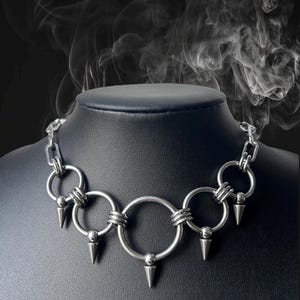 Peut inclure: Un collier ras du cou argenté composé d'une série de grands anneaux reliés par de petites chaînes. Chaque grand anneau est orné d'un pendentif en forme de pointe argentée. Le collier est présenté sur un mannequin noir.