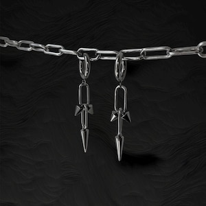 Op de afbeelding: Twee zilverkleurige oorbellen met een kettingontwerp. Elke oorbel heeft een kleine, puntige bedel en een lange, puntige hanger. De oorbellen hangen aan een zilveren ketting tegen een donkere, getextureerde achtergrond.