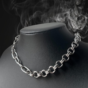 Op de afbeelding: Een zilverkleurige ketting met verschillende schakelvormen, gepresenteerd op een zwarte mannequin. De ketting heeft een mix van ovale en cirkelvormige schakels, wat zorgt voor een gedurfde en moderne uitstraling. De achtergrond is donker met een rokerig effect.