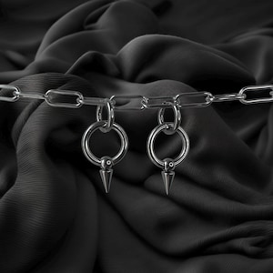 Puede incluir: Una cadena plateada con dos colgantes circulares, cada uno con un anillo y un cono que apunta hacia abajo. Los colgantes están conectados a la cadena. El fondo es una tela oscura y texturizada.
