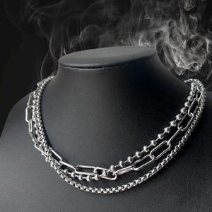 Może przedstawiać: Srebrny zestaw naszyjników na czarnym manekinie. Naszyjnik składa się z trzech warstw: łańcuszka z małych koralików, łańcuszka z prostokątnymi ogniwami i łańcuszka z małymi, kwadratowymi ogniwami. Tło jest ciemne z efektem dymu.