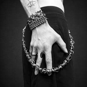 May include: A monochrome image featuring metal jewellery. A spiked bracelet and a chainmail bracelet are on the wrist. A chainmail necklace is held in the hand. The jewellery has a gothic or punk aesthetic.