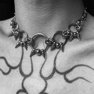 Może przedstawiać: Srebrny naszyjnik typu choker w stylu punk rock. Naszyjnik posiada trzy duże pierścienie połączone ogniwami łańcucha, każdy ozdobiony stożkowymi kolcami i małymi metalowymi koralikami. Projekt jest odważny i ostry.