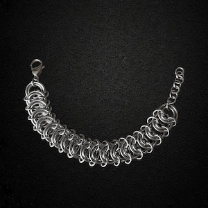 Puede incluir: Una pulsera de cota de malla en tono plateado construida con anillos de metal entrelazados. La pulsera cuenta con un cierre de mosquetón en un extremo y una cadena de extensión corta con eslabones circulares más pequeños en el otro.
