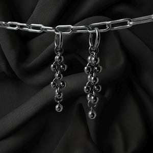 May include: A pair of silver-toned earrings with a cluster of small, round beads hanging from a hoop. The earrings are suspended against a dark, textured background, with a silver chain visible at the top.