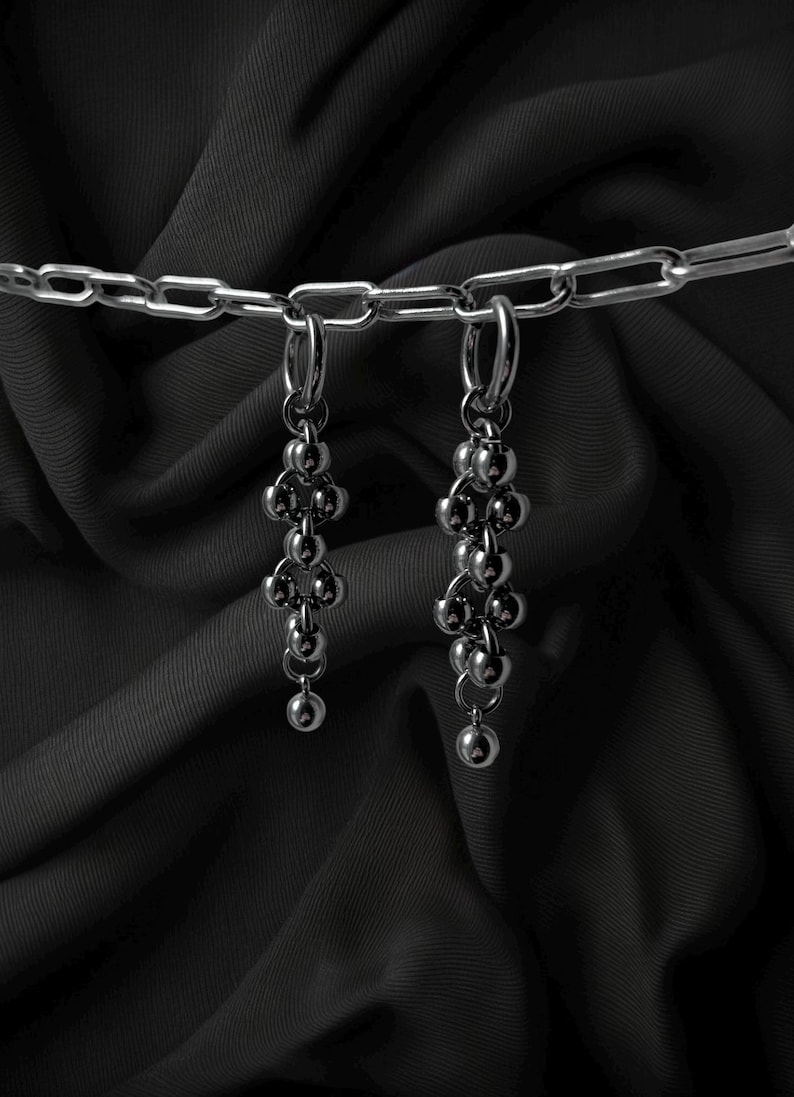May include: A pair of silver-toned earrings with a cluster of small, round beads hanging from a hoop. The earrings are suspended against a dark, textured background, with a silver chain visible at the top.