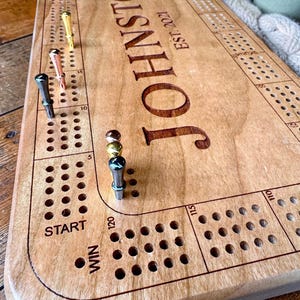 Peut inclure: Un plateau de cribbage rectangulaire en bois clair, gravé avec le texte JOHNSTON EST. 2021. Le plateau présente des rangées de trous percés pour le jeu, marqués START et WIN, accompagnés de six pions métalliques dorés, cuivrés et argentés.
