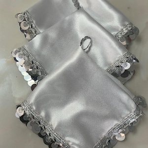 Puede incluir: Tres servilletas cuadradas de satén plateado. Cada servilleta tiene un borde decorativo de lentejuelas y adornos plateados. Pequeños anillos plateados se colocan encima de las servilletas.