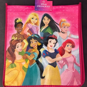 May include: Pink tote bag featuring multiple Disney Princesses. The bag has a pink border and a tag that reads "Disney Princess". The princesses include Tiana, Rapunzel, Mulan, Cinderella, Belle, Jasmine, Snow White, and Ariel.