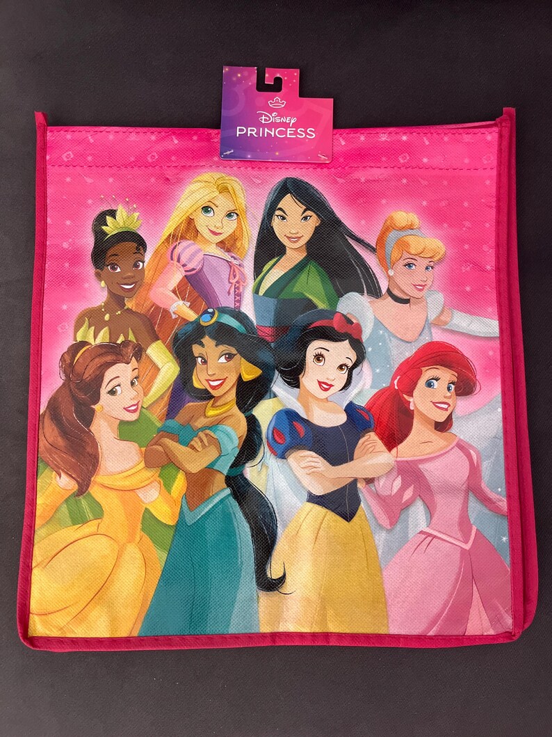 May include: Pink tote bag featuring multiple Disney Princesses. The bag has a pink border and a tag that reads "Disney Princess". The princesses include Tiana, Rapunzel, Mulan, Cinderella, Belle, Jasmine, Snow White, and Ariel.