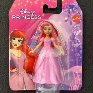 Puede incluir: Mu&ntilde;eca Disney Princess con vestido rosa y pelo rojo, empaquetada en una tarjeta rosa y morada. El embalaje presenta el logotipo de Disney Princess y una ilustraci&oacute;n del personaje. La mu&ntilde;eca es apta para mayores de 3 a&ntilde;os.
