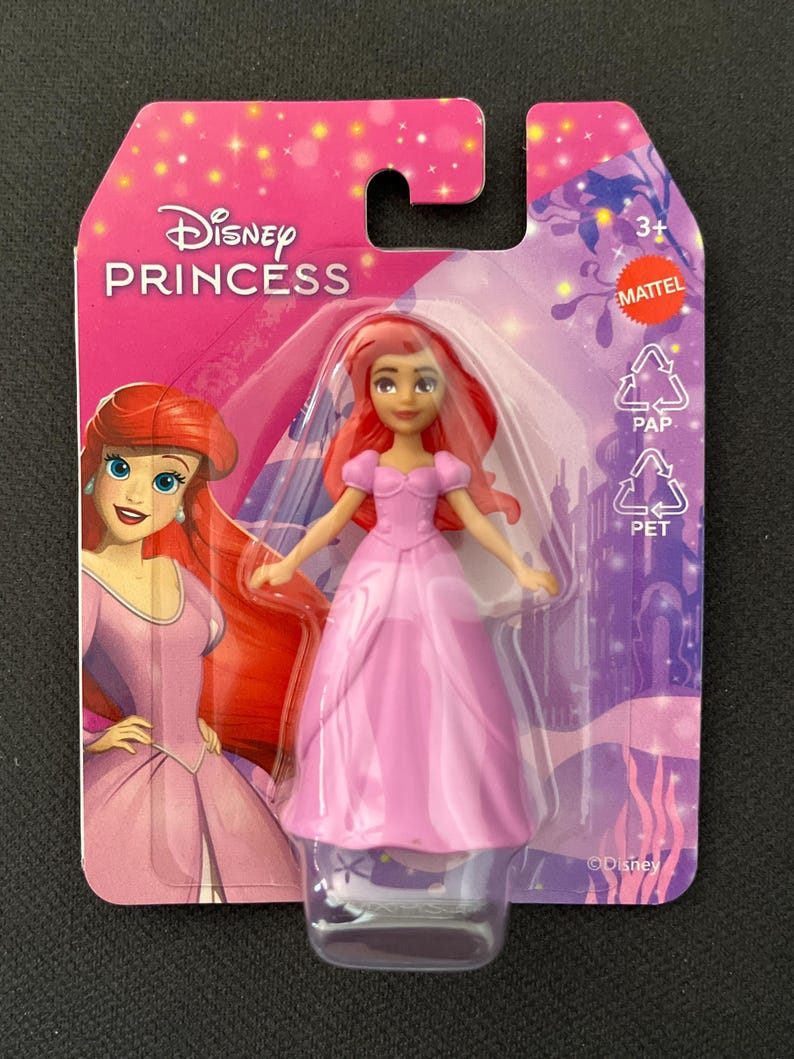 Puede incluir: Mu&ntilde;eca Disney Princess con vestido rosa y pelo rojo, empaquetada en una tarjeta rosa y morada. El embalaje presenta el logotipo de Disney Princess y una ilustraci&oacute;n del personaje. La mu&ntilde;eca es apta para mayores de 3 a&ntilde;os.