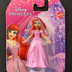 Puede incluir: Mu&ntilde;eca Ariel de Disney Princess con un vestido rosa, empaquetada en una tarjeta rosa y morada. La tarjeta presenta el logotipo de Disney Princess y una ilustraci&oacute;n de Ariel. La mu&ntilde;eca est&aacute; en un soporte de pl&aacute;stico transparente.
