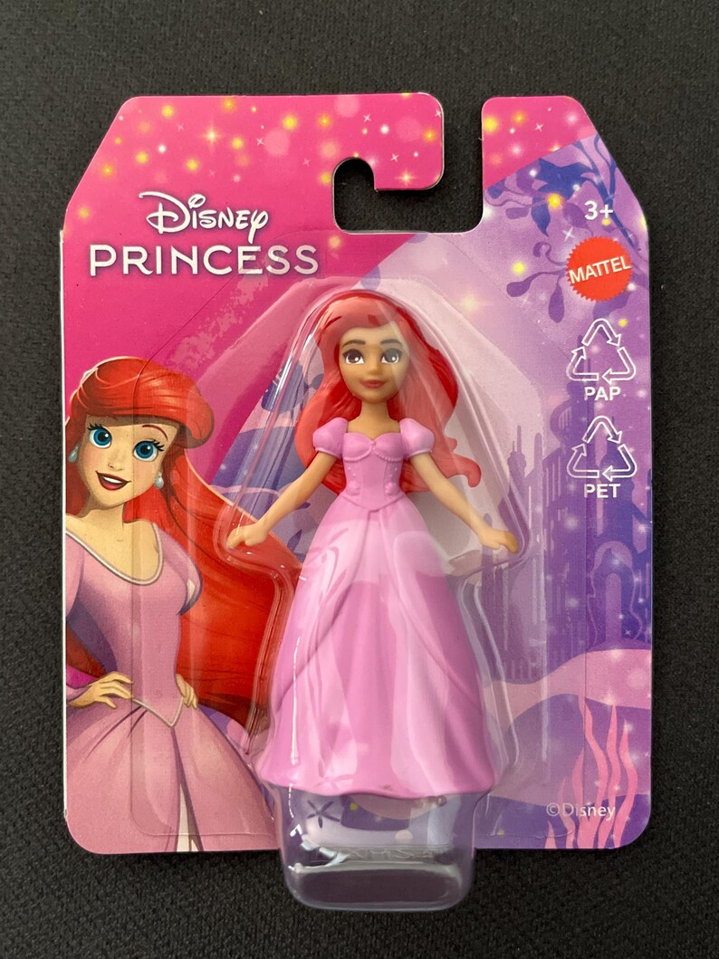Puede incluir: Mu&ntilde;eca Ariel de Disney Princess con un vestido rosa, empaquetada en una tarjeta rosa y morada. La tarjeta presenta el logotipo de Disney Princess y una ilustraci&oacute;n de Ariel. La mu&ntilde;eca est&aacute; en un soporte de pl&aacute;stico transparente.