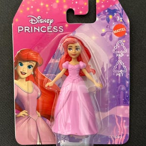 Puede incluir: Mu&ntilde;eca Ariel de Disney Princess con un vestido rosa, empaquetada en una tarjeta rosa y morada. La tarjeta presenta el logotipo de Disney Princess y una ilustraci&oacute;n de Ariel. La mu&ntilde;eca est&aacute; en una funda de pl&aacute;stico transparente.