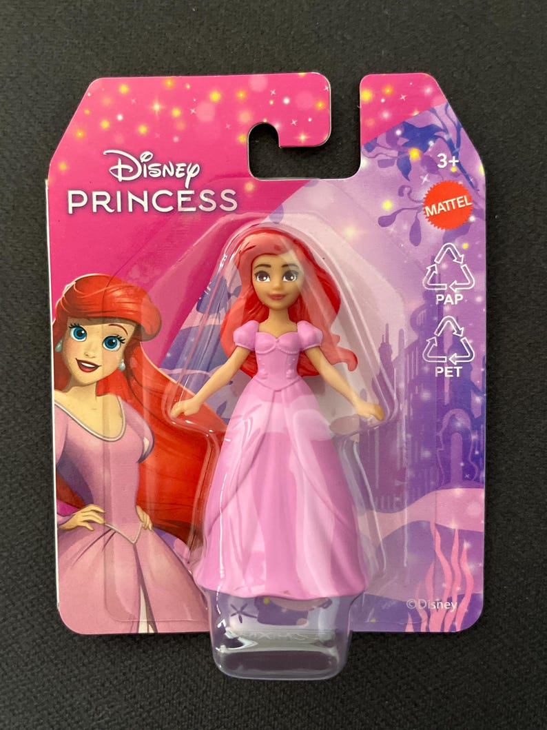 Puede incluir: Mu&ntilde;eca Ariel de Disney Princess con un vestido rosa, empaquetada en una tarjeta rosa y morada. La tarjeta presenta el logotipo de Disney Princess y una ilustraci&oacute;n de Ariel. La mu&ntilde;eca est&aacute; en una funda de pl&aacute;stico transparente.