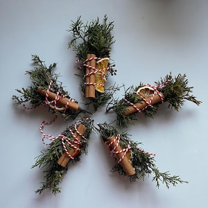 May include: Five bundles of dried greenery, cinnamon sticks, and dried orange slices, tied with red and white twine. The arrangement is set against a light background, showcasing the natural colors and textures of the components.