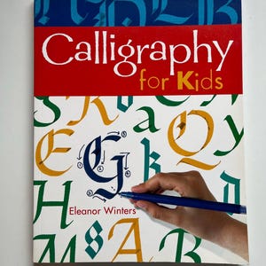 May include: A book titled "Calligraphy for Kids" with a red and blue cover. The cover features the title in white letters and colorful calligraphy examples. A hand is holding a blue pen, demonstrating calligraphy.