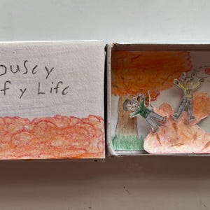 May include: A rectangular cardboard matchbox featuring a hand-drawn scene of two mouse-like figures playing in orange autumn leaves. The exterior cover displays the handwritten text "Mousy Leafy Life" above a pile of orange leaves in colored pencil.