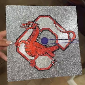 May include: A silver glitter graduation cap with a red and white dragon design. The dragon is in a hexagonal shape with a blue circle in the center.