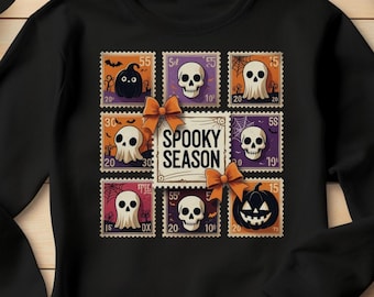 May include: Black crewneck sweatshirt featuring a Halloween-themed design. The design includes postage stamp-style graphics with ghosts, skulls, and a pumpkin, along with the text "Spooky Season". A black circle with the word "Black" is in the upper left corner.