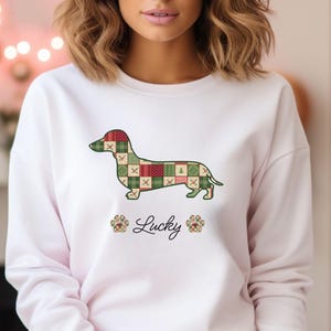 Op de afbeelding: Witte sweatshirt met een teckel hond ontwerp met een patchwork patroon in rood, groen en crème. Het woord "Lucky" staat in cursief onder de hond, met pootafdrukken aan weerszijden. De trui heeft een roze rand.