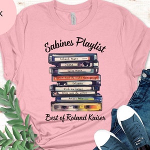 May include: Pink t-shirt with a graphic of a stack of vintage cassette tapes with the text "Sabines Playlist" and "Best of Roland Kaiser".