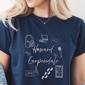 Op de afbeelding: Marineblauwe t-shirt met witte lijntekening van een koptelefoon, platenspeler, lippen, een microfoon, een cassettebandje, voetstappen en een deur. De tekst "Howard Carpendale &" is cursief geschreven.