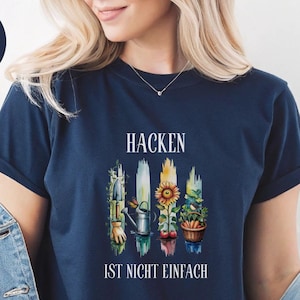 Könnte beinhalten: Marineblaues T-Shirt mit dem Text "HACKEN IST NICHT EINFACH" und einem Aquarelldesign von Gartengeräten, einer Sonnenblume und Gemüse. Das Shirt hat kurze Ärmel und einen Rundhalsausschnitt.