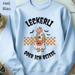 Pode incluir: Camisola azul clara com um design de Halloween. O design apresenta um cão vestido de fantasma, segurando uma abóbora, com o texto alemão "LECKERLI ODER ICH BEISSE" e morcegos. A camisola tem gola redonda e mangas compridas.