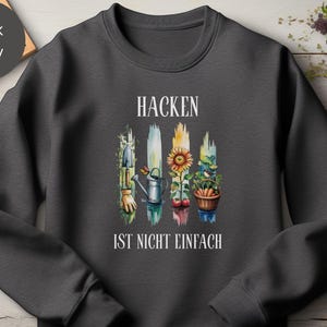 Könnte beinhalten: Dunkelgraues Sweatshirt mit einem Aquarelldesign mit Gartengeräten, einer Sonnenblume und einem Korb mit Gemüse. Der Text "HACKEN IST NICHT EINFACH" ist in Weiß gedruckt. Das Sweatshirt ist aus weichem Material.