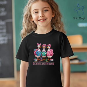 Peut inclure: T-shirt noir avec un motif de trois oies portant des chapeaux et des sacs à dos, avec le texte "Endlich erstklassig" en rose. Les oies sont vêtues de tenues bleues, vertes et roses. Le t-shirt est porté par une jeune fille.