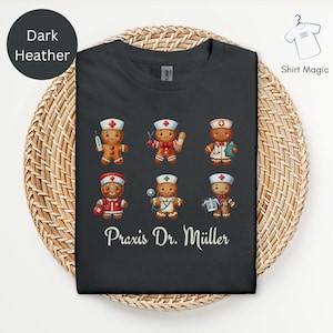 Puede incluir: Camiseta de color gris oscuro con personajes de médicos de pan de jengibre con uniformes médicos y herramientas. El texto "Praxis Dr. Müller" está impreso debajo. La camiseta se muestra en una cesta tejida.