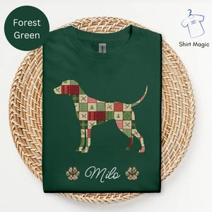 Könnte beinhalten: Forstgrünes T-Shirt mit einer Hundesilhouette, gefüllt mit einem Patchwork-Design in Rot, Grün und Creme. Der Name "Milo" steht in Schreibschrift unter dem Hund, mit Pfotenabdrücken auf beiden Seiten. Das Shirt wird auf einem geflochtenen Korb präsentiert.
