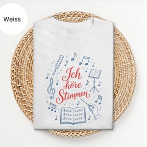 Peut inclure: Un t-shirt blanc avec un texte central en écriture rouge indiquant Ich höre Stimmen, entouré d'illustrations bleues de notes de musique, d'une clé de sol, d'un diapason, d'un pupitre, d'une partition ouverte et d'une main tenant une baguette.