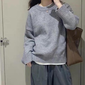 Könnte beinhalten: Ein grauer Strickpullover mit langen Ärmeln und Rundhalsausschnitt. Der Pullover wird mit einer weiten grauen Hose und einer braunen Ledertasche kombiniert.