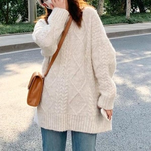 Peut inclure: Un pull blanc en tricot torsadé à la coupe ample et aux manches longues. Le pull est porté avec un jean bleu et des chaussures noires.