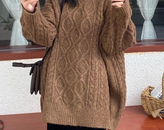 May include: A brown cable-knit sweater with a round neckline. The sweater is paired with a black midi skirt. A brown shoulder bag and black shoes complete the outfit. A small basket with a handle sits on a wooden surface.