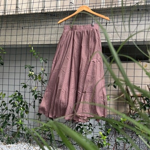 Op de afbeelding: Een stoffige roze rok die aan een houten hanger hangt. De rok is gemaakt van een zachte, vloeiende stof en heeft een geplooide taille.