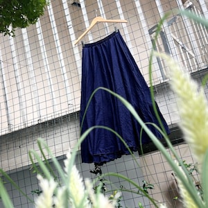 Op de afbeelding: Een donkerblauwe maxi-rok die aan een houten hanger hangt tegen een wit metalen hek. De rok is gemaakt van een zachte, vloeiende stof en heeft een eenvoudig, elegant ontwerp.