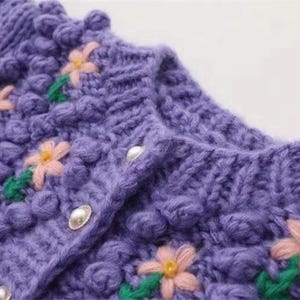 Puede incluir: C&aacute;rdigan de punto morado con una superficie texturizada y esponjosa. El c&aacute;rdigan presenta flores bordadas en rosa, amarillo y verde, y botones perlados.