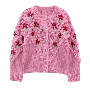 Puede incluir: Un c&aacute;rdigan de punto rosa con un dise&ntilde;o bordado floral. El c&aacute;rdigan tiene cierre de botones y un punto texturizado y abultado.