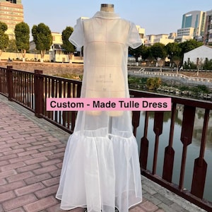 May include: A white tulle dress with a ruffled hem. The dress is on a mannequin and is displayed against a backdrop of a city skyline and a river.