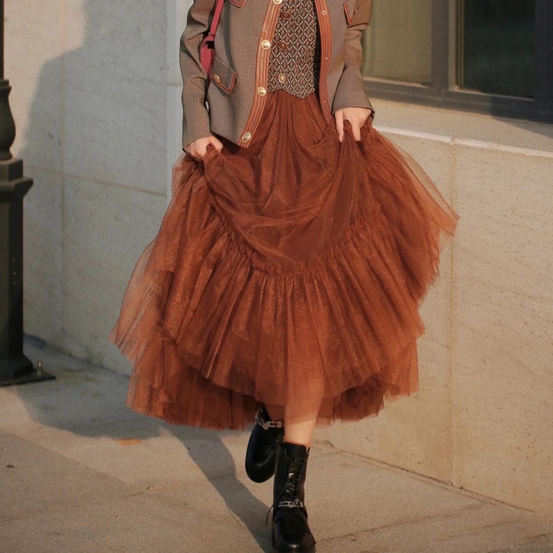 May include: A brown tulle skirt with multiple layers and a ruffled hem. The skirt is worn with a brown jacket and black boots.