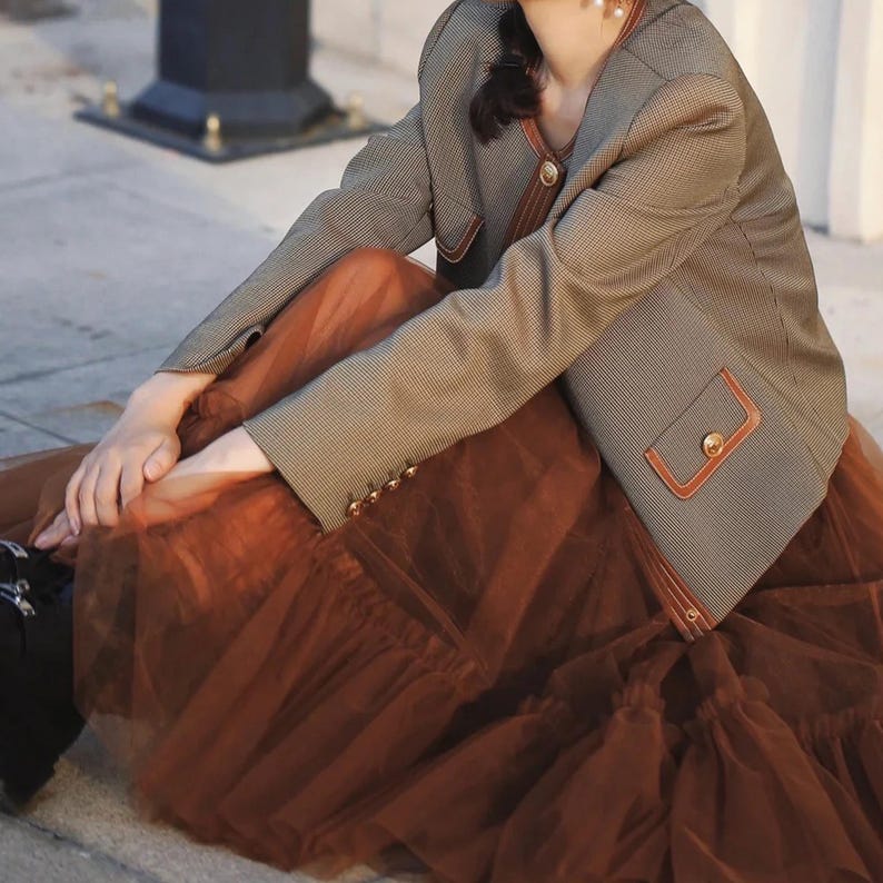 May include: A brown tulle skirt with a brown and white plaid blazer. The blazer has a brown leather trim and gold buttons.